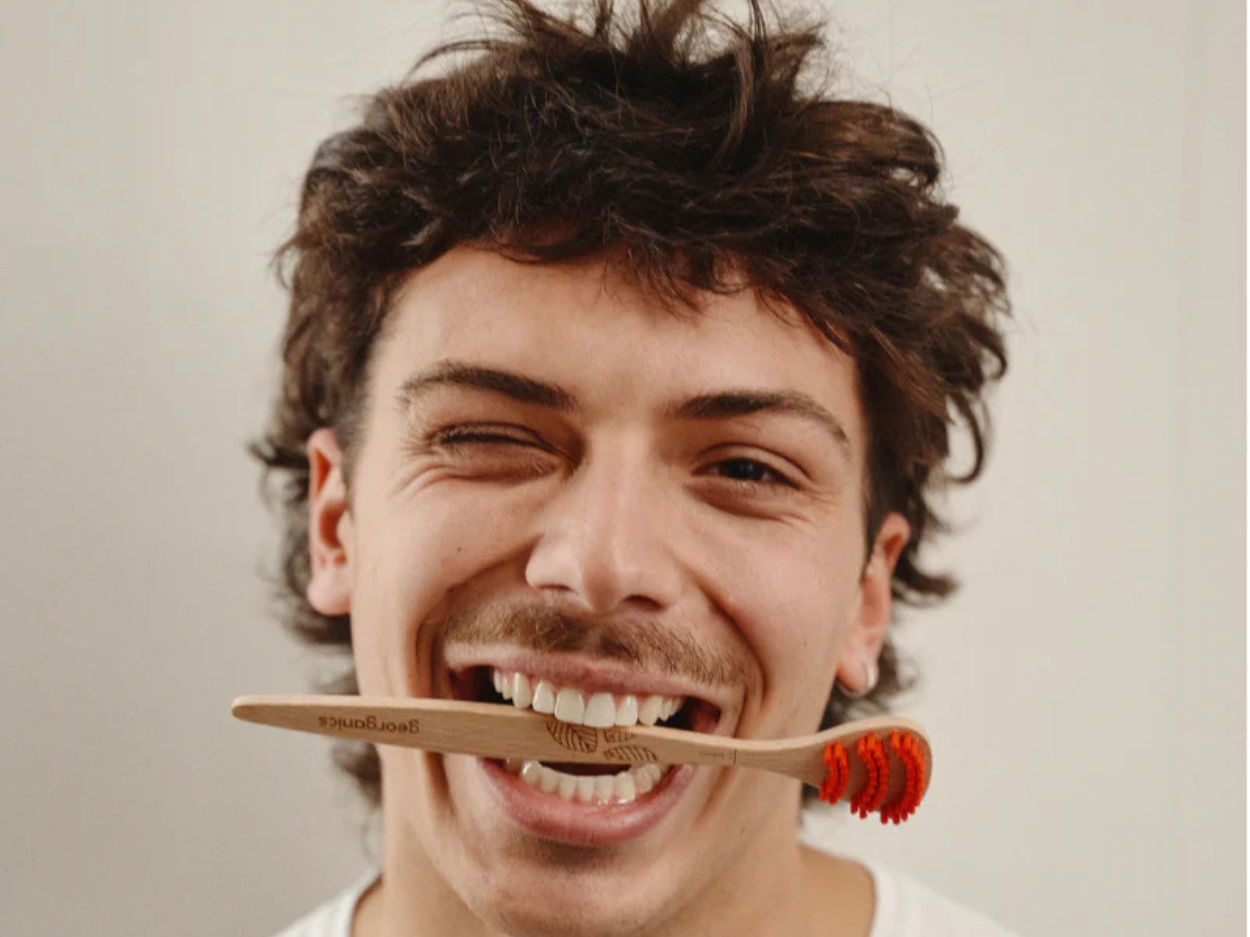 Smiling man holding a wooden Georganics tongue brush between his teeth.